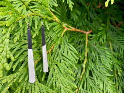 Two earrings are hanging off of lush green evergreen branches. They are long black dangles that have been dipped in silver metallic paint. 