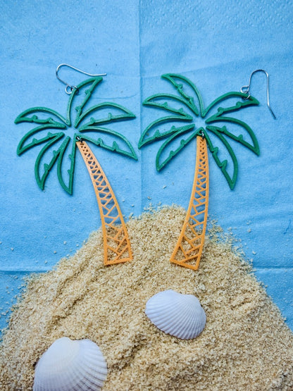 In front of a bright sky blue background is a mound of sand like an island. There are white shells in the sand. At the top of the mound are two 3D printed R+D earrings that are shaped like palm trees. The fronds are kelly green and burst out in a lively way. The trunks are orange and curve to the swaying top.