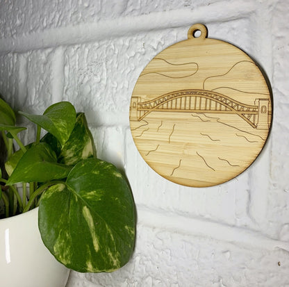 Hanging on a white brick wall and next to a houseplant is a laser cut wall hanging. It is cut from bamboo, a fast growing and sustainable grass. Etched in it a picture of the Hell Gate bridge in Astoria. This bridge is an icon of the area and looked on from Asttoria Park. This can be used as a ornament in the holidays or a wall hanging all year round. It can be painted like a paint by number or left plain.