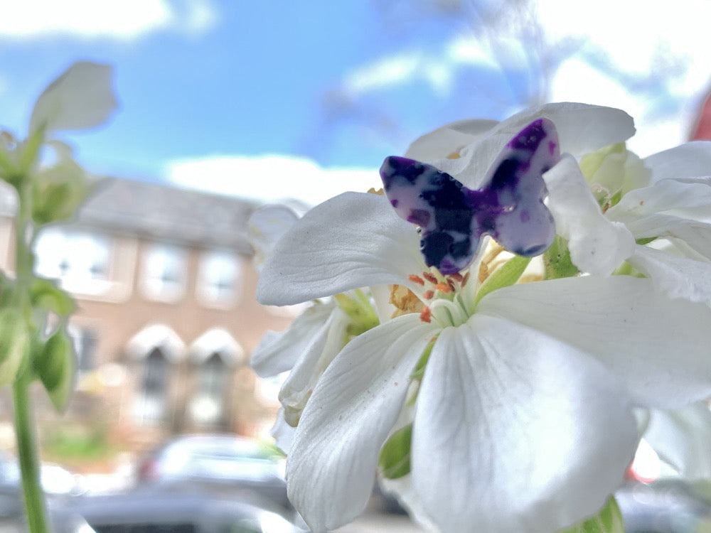In front of a bright blue sky and resting on a white flower is a cast ring in the shape of a butterfly. It is cast from recycled 3D prints in hues of purple, white, and black. It has the appearance of marble or granite. 