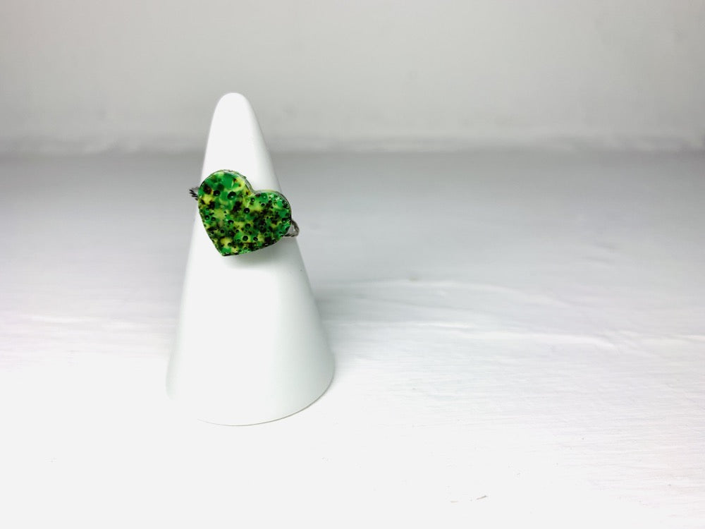 On a white background and slipped on a white cone ring holder is a ring cast from recycled 3D prints. The ring has a heart charm and a thin twisted metal band. The charm is in the shape of a heart and uses various shades of green. 