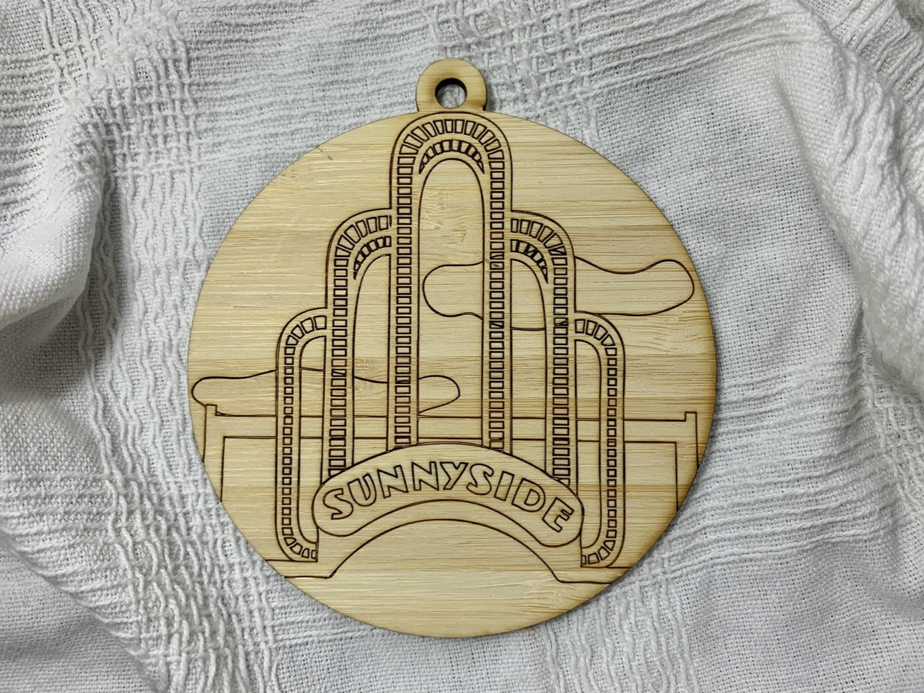 Shown on a white fabric background is a laser cut wall hanging. It is cut from bamboo, a fast growing and sustainable grass. Etched in it a picture of the iconic Sunnyside Arch in the center of the Queens neighborhood. Each piece can be painted like a paint by number or left plain. It can hang from your Christmas Tree or on you wall all year round.