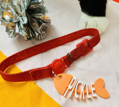 A silver foil cat toy is in the upper corner and you can see a black and white cat's paw standing in front of a red collar with a tag on it. There is a cat tag that is shaped like a cartoon fish that has been eaten. The head, tail, and backbone is bright orange. Where the bones would be, there are letters to form the name Pickle in white.