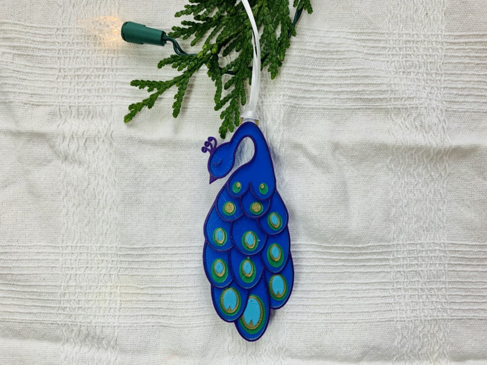 On a white fabric background and hanging off of a evergreen spring is a R+D 3D printed  peacock ornament. It has a blue body, with purple outlines. On the long elegant feathers are green, gold and teal accents.