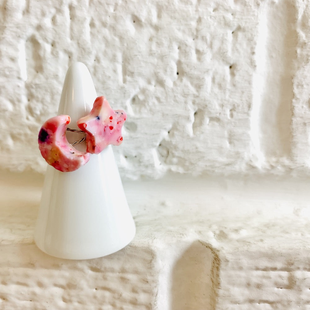 On painted white bricks is a white ring holder with two cast rings on it. One ring is shaped like a moon, the other a star. They are cast from recycled 3D printed pieces in various warm and pink hues. 