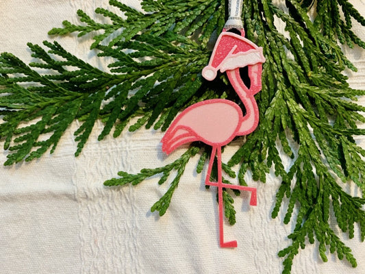 On a white fabric background with sprigs of evergreen is a R+D 3D printed ornament. It is a bright pink flamingo that has one leg up and is wearing a hot pink santa hat. 