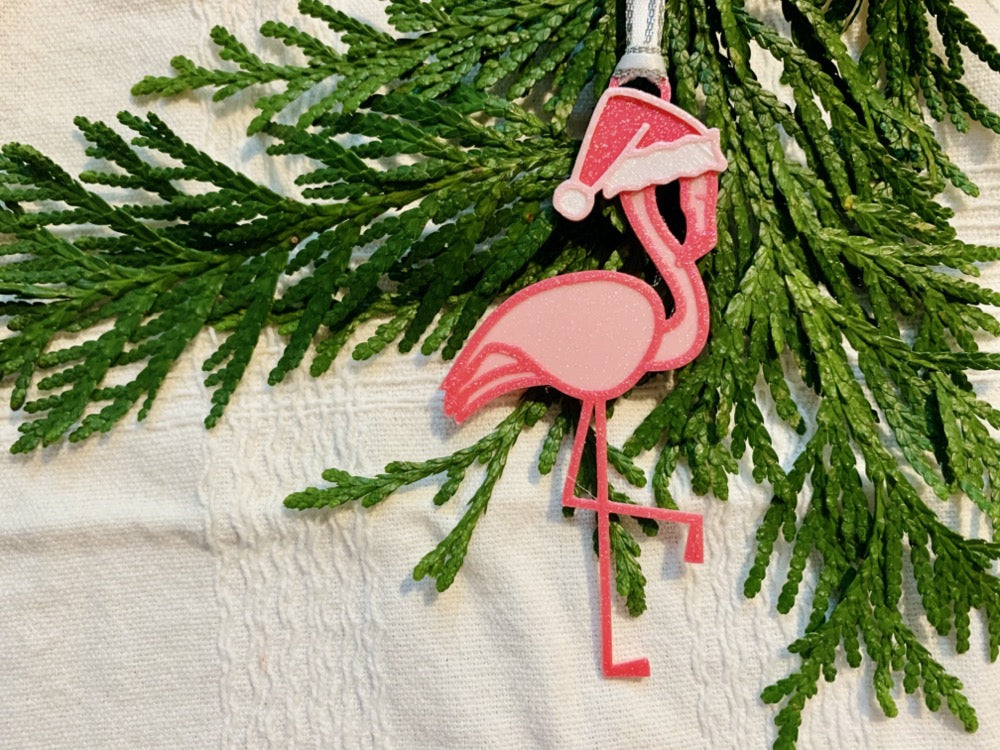 On a white fabric background with sprigs of evergreen is a R+D 3D printed ornament. It is a bright pink flamingo that has one leg up and is wearing a hot pink santa hat. 
