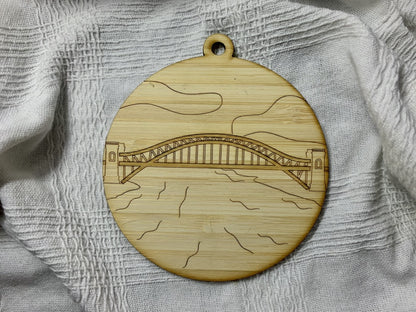 Shown on a white fabric background is a laser cut wall hanging. It is cut from bamboo, a fast growing and sustainable grass. Etched in it a picture of the Hell Gate bridge in Astoria. This bridge is an icon of the area and looked on from Asttoria Park. This can be used as a ornament in the holidays or a wall hanging all year round. It can be painted like a paint by number or left plain. 