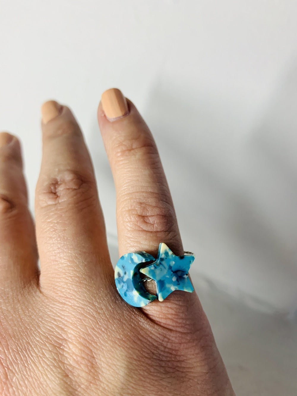 In front of a white background is someone's hand with two rings on their pinky finger. The rings are made from recycled 3D prints with teal, blue, white, and black colors intermixed to be speckled like granite.