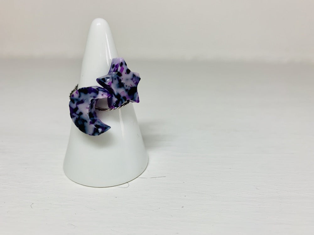 On a white background is a ring holder with two cast rings resting on it. One ring is a present moon shape, the other is a star. Both have a twisted metal band. The ring is cast from recycled 3D prints in shades of purple, black and white to create a marbled or granite like appearance.