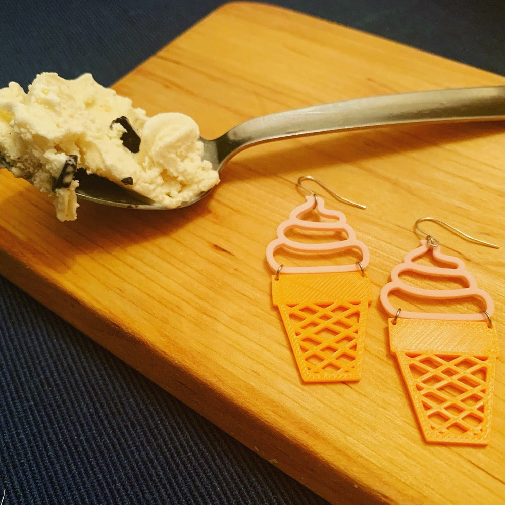 Two ice cream cone earrings are laying on a wooden cutting board next to a spoon piled with ice cream. The earrings have pink tops swirled like soft serve and orange bottoms that look like a sugar cone. 