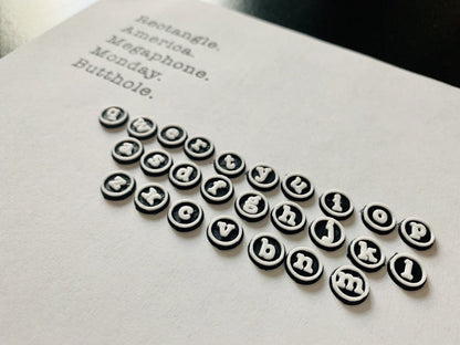 In the foreground are 26 earrings. They are shaped as typwriter keys: circles with white outlines and a lowercase letterin the center. The earrings are arranged in the order of a standard keyboard. They are all 3D printed and laying on a piece of white printer paper. In thr far background are 5 words: rectangle, america, megaphone, monday, and butthole. In Parks and Recreation, these are the words Ron Swanson types after getting a typewriter and saying he will type every word he knows. 