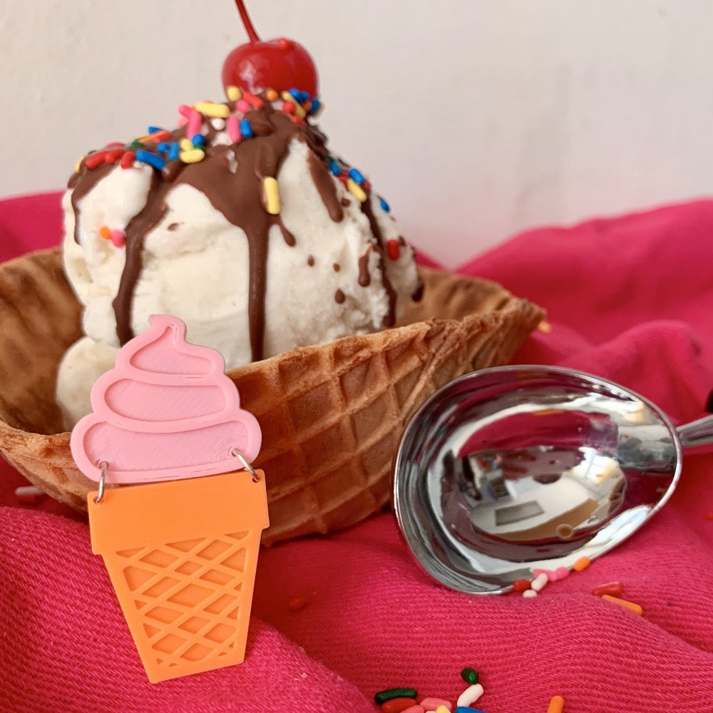 A plant based 3D Printed pin shaped like an ice cream cone is in the foreground. It has a pink swirled soft serve top and an orange sugar cone base. In the background is a ice cream sundae in a waffle cone bowl with vanilla ice cream, chocolate sauce, sprinkles, and a bright red cherry on top. An ice cream scoop rests next to it all.
