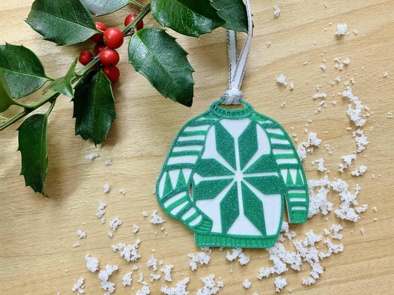 Shown on a wood shelf is a sprig of holly and some white snow. Laying across this is a 3D printed ornament from R+D. It is in the shape of a chunky sweater. This particular one is white and green and has stripes, trees on the sleeves, and a snowflake in the center.