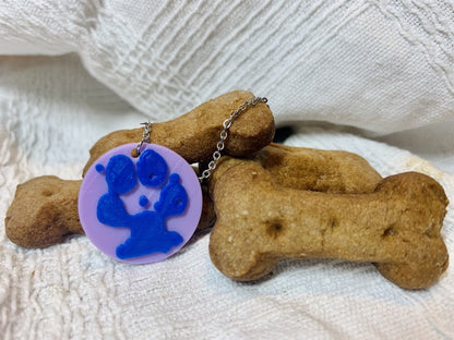 Shown on white fabric is a pile of dog bone treats. In front of that is a 3D printed pendant. The pendant is a 1 inch circle that is light purple. Printed on the pendant is a blue paw print that is customized. 