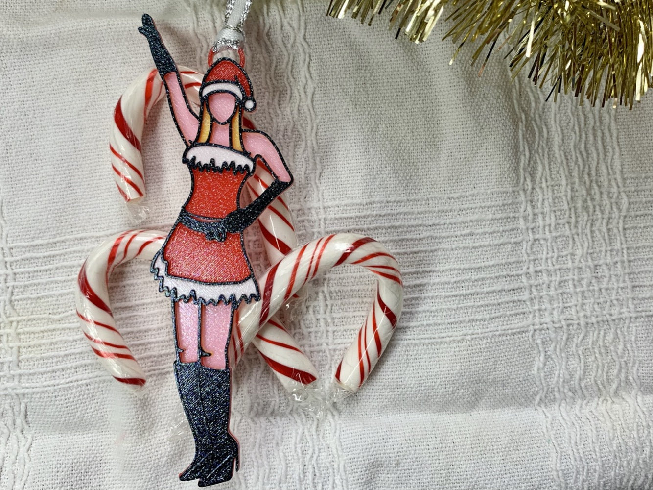 On a white fabric background there is a bit of gold garland and candy canes. Laying on top of these is a 3D printed ornament from R+D. The ornament is printed in a plant based filament. It is shaped like Regina George from the movie Mean Girls. She is striking the iconic pose at the beginning of performing Jingle Bell Rock, wearing black gloves and boots and a red outfit with white trim. The entire ornament is covered in glitter to shimmer and shine in the light. 