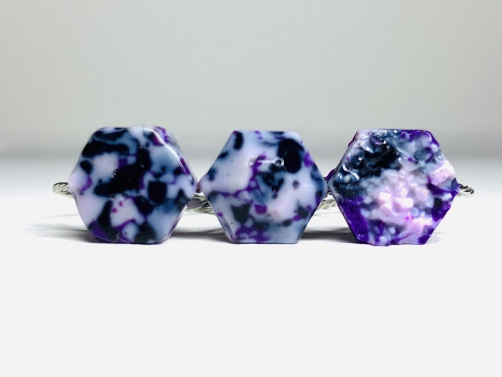 On a bright white background are three rings. They are all cast using recycled 3D prints. They have a speckled or granite like appearance with shades of white, black, purple, and light purple plant based filaments. They are hexagons in shape; one has a smooth surface, another a faceted surface, and finally a bumpy geode surface. 