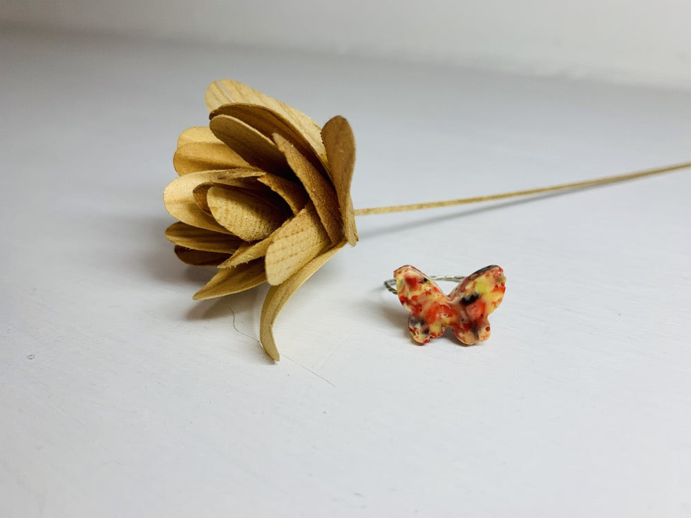 On a white background is a ring that is cast from recycled 3D prints. The ring is in a butterfly shape with red, yellow, orange, and black colors intermixed to be speckled like granite and resting next to a small wooden flower. 