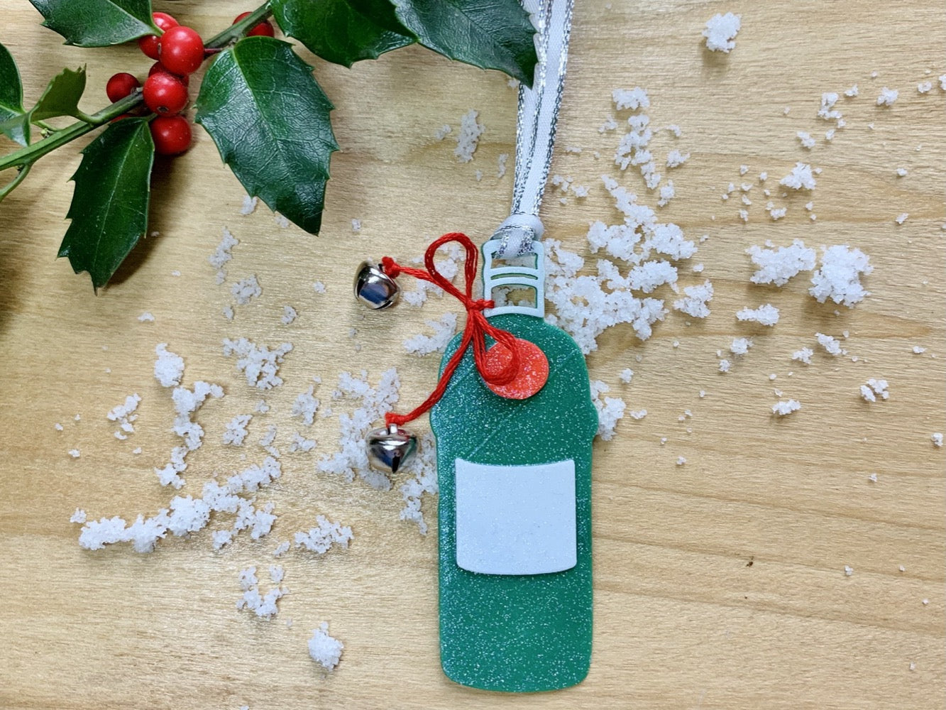 Shown on a wood background with a sprig of holly and white snow is a 3D printed R+D ornament. It is in the shape of a gin bottle: green with a white label and a red emblem. Tied around the neck is a red bow that has two jingle bells attached. The entire ornament is covered in glitter to give it a shimmer and shine.
