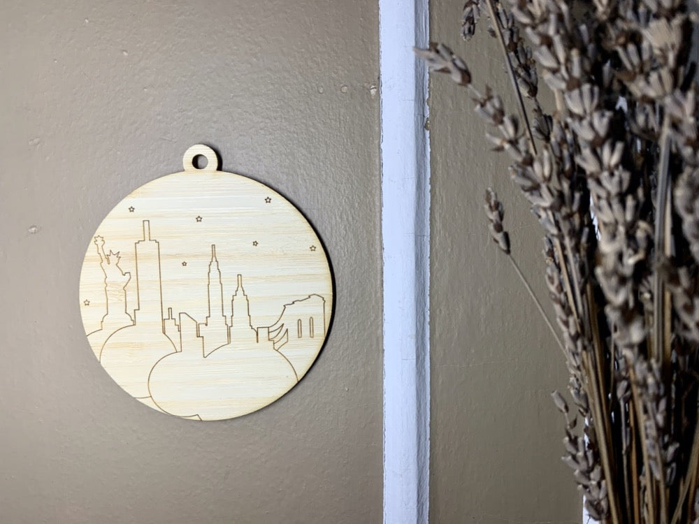 Shown on a brown wall with drip lavender to the side is a laser cut wall hanging. It is cut from bamboo, a fast growing and sustainable grass. Etched in it a picture of the New York City Skyline with the Statue of Liberty, Empire State Building, Chrysler Building and Brooklyn Bridge all visible. Each piece can be painted like a paint by number or left plain. It can hang from your Christmas Tree or on you wall all year round.