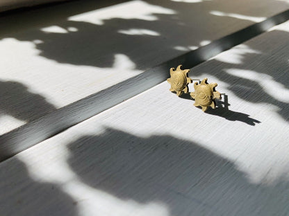 Shown in the bright afternoon sun are two gold 3D printed earrings shaped like suns. They are in a sun beam, but surrounded by shadows casting from plants.