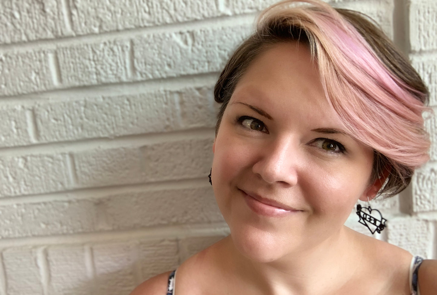 Rebekah Thornhill, the owner and maker behind R+D is shown in front of a white brick wall. One 3D printed earring is visible, it is in the shape of a heart tattoo: a heart, with  a banner twisting around it and two roses. The banner says R+D on it and can be customized to include any word. 