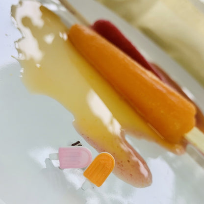 In the foreground are two R+D studs shaped like popsicles. They are complementary colors, one is light pink, the other is orange, and they both have white sticks. In the background, there  are two melting popsicles one is orange and one is dark red.