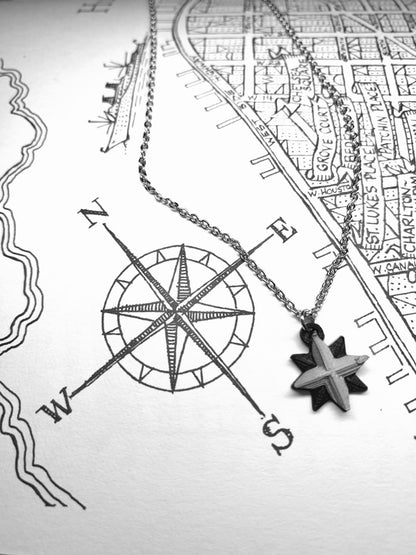 Laying across a drawn map and compass is an R+D necklace. The necklace is black and silver and shaped like a compass with 8 points. 
