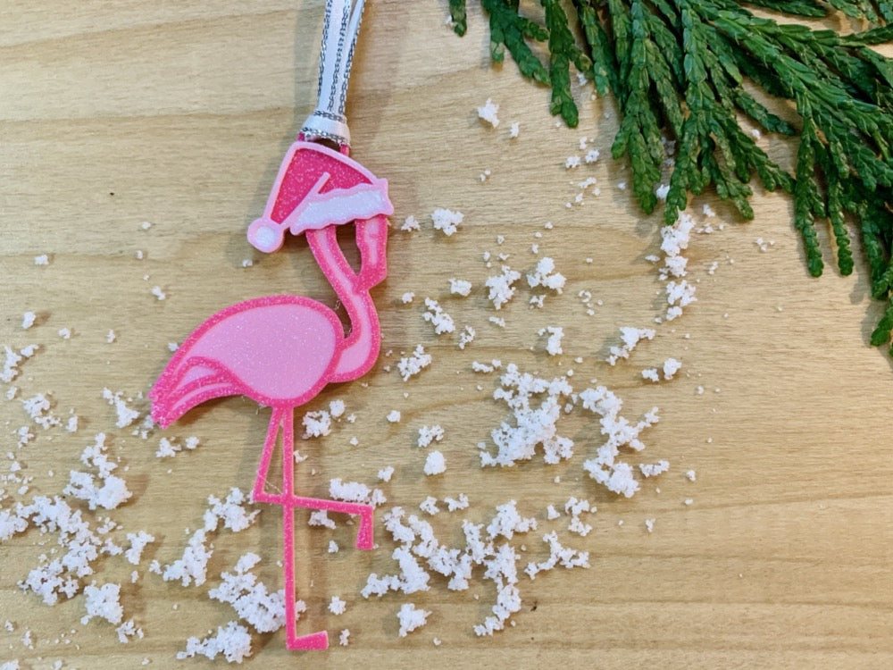 On a wooden shelf with sprigs of evergreen and white snow is a R+D 3D printed ornament. It is a bright pink flamingo that has one leg up and is wearing a hot pink santa hat.