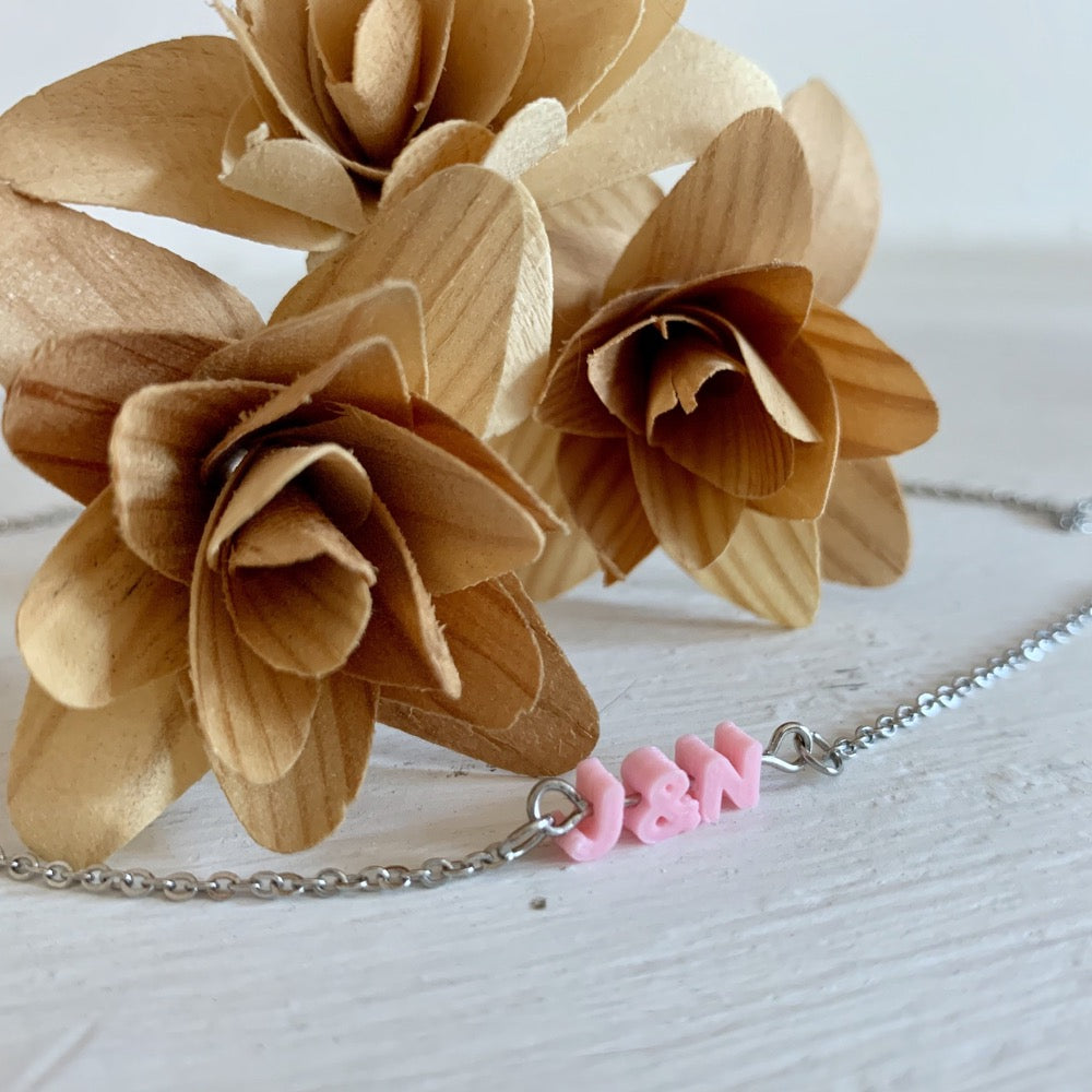 In front of three wooden flowers is a necklace with small letters on a metal bar. The letters are like beads and will rotate and move around. This necklace has 3 characters (J&N) in a light pink. 