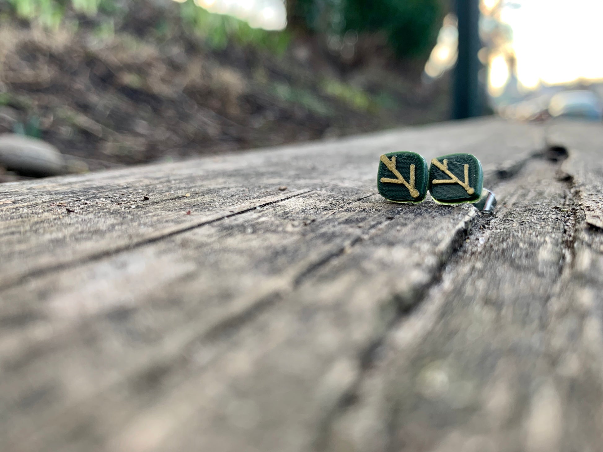 In focus are two R+D earrings shaped like leaves. They are olive green with gold veins. They are resting on weathered wood planks and there is a park blurry in the background.