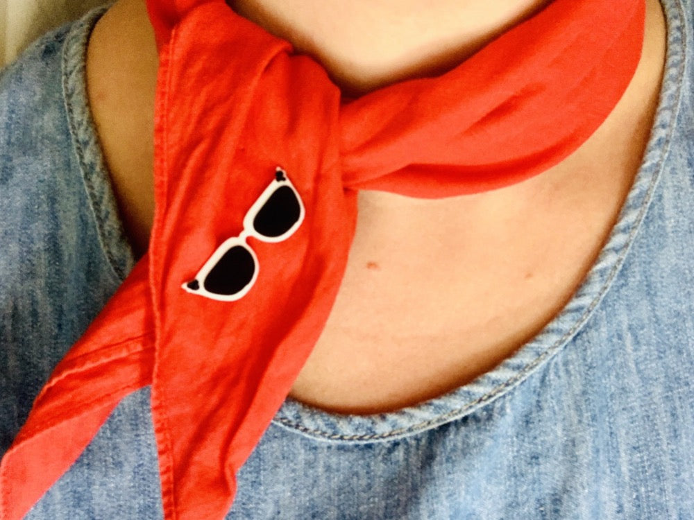 A red scarf is around a person's neck and twisted to one side. Holding the scarf in place is a 3D printed pin from R+D shaped like cat eye sunglasses. They are white  frames, with black lenses and black dots in the upper corners of the frames. 