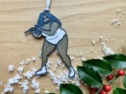 On a light wood shelf is a sprig of holly and a scattering of snow. Resting on top of this is a R+D 3D printed ornament. The ornament is shaped like the singer, rapper, and musician Lizzo. She is wearing a white leotard, bend slightly, and playing her flute name Sasha Flute. The entire ornament is printed with a plant based filament and covered in glitter to give it a perfect shimmer and shine. 