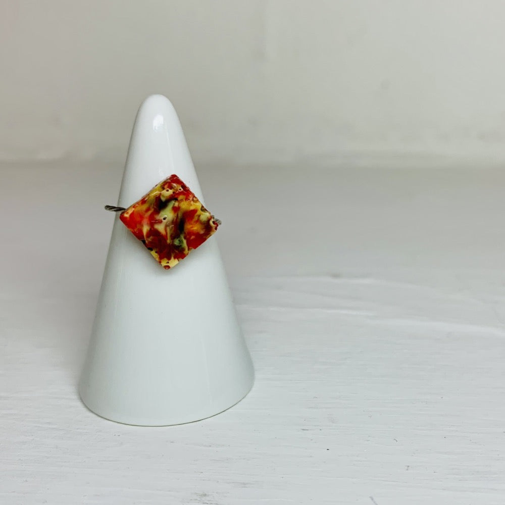 On a white background and slipped on a white cone ring holder is a ring that is cast from recycled 3D prints. The ring is in the shape of a faceted square with red, yellow, orange, and black colors intermixed to be speckled like granite.