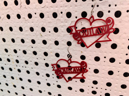 Two earrings are hanging off of a background with white paint and holes across it. The earrings are a dark merlot red and shaped like heart tattoos--they are hearts with a banner twisting around it and two roses. The banner reads outlaw, but can be personalized to any word. They are 3D printed in an eco friendly filament.