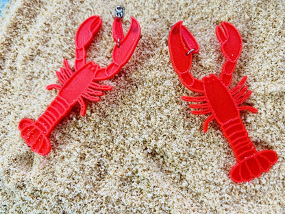 Shown on a bed of sand are two 3D printed R+D earrings. The earrings are bright red and shaped like lobsters. The larger crusher claw on the lobster wraps around the earlobe when being worn.