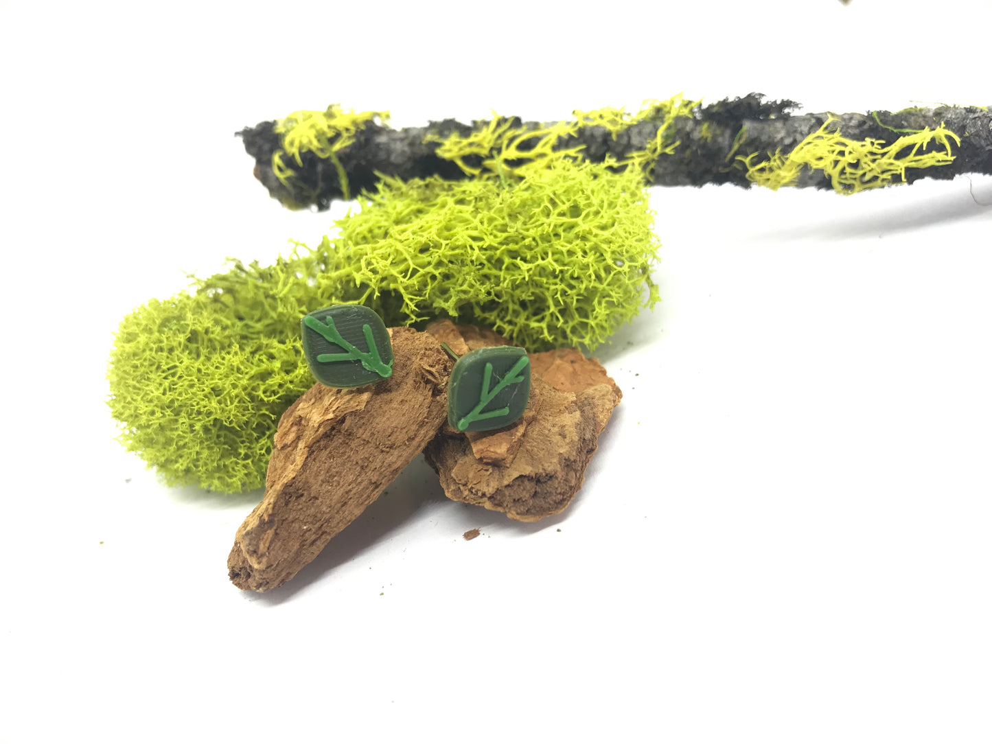 In a stark white frame, there is a stick, moss, and small pieces of wood. Resting on the wood are two R+D earring studs that are shaped like leaves. They are dark olive green with kelly green veins.