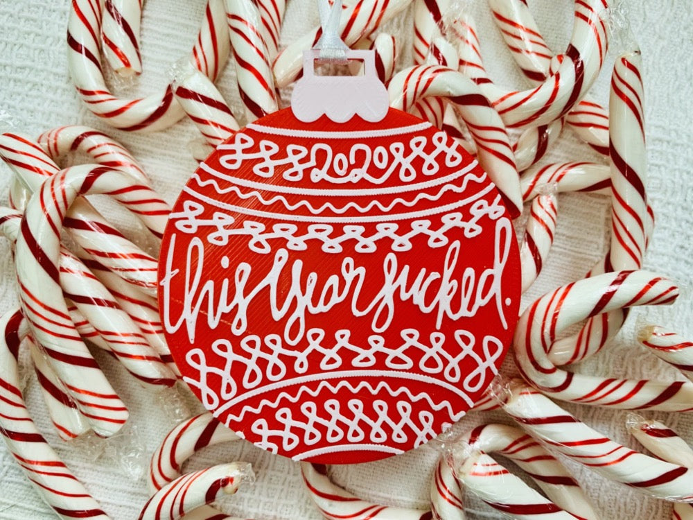 On a white fabric background there are lots of red and white peppermint candy canes and a R+D 3D printed ornament. The ornament is shaped like a traditional bulb. It is bright red with white drawings and script on it. Worked into the doodles are the year 2020 and then the words, "this year sucked".