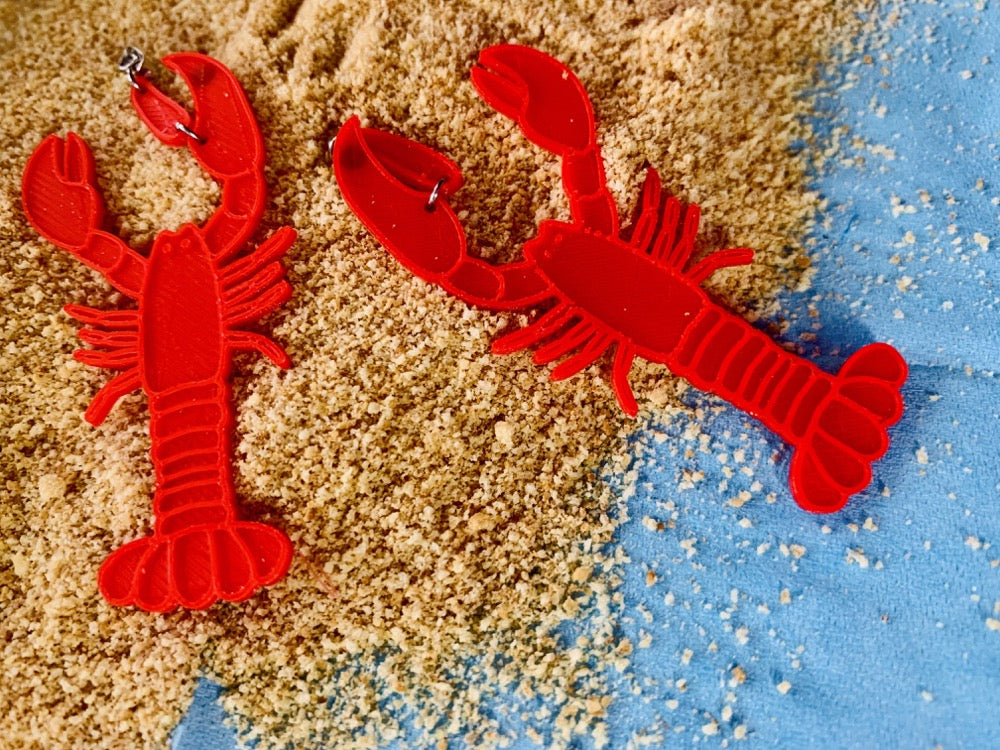 A baby blue background is covered part way with mounds of crisp tan sand. Laying on top are two R+D earrings that are shaped like lobsters. They are bright red and the larger crusher claw is where the stud wraps around the earlobe.