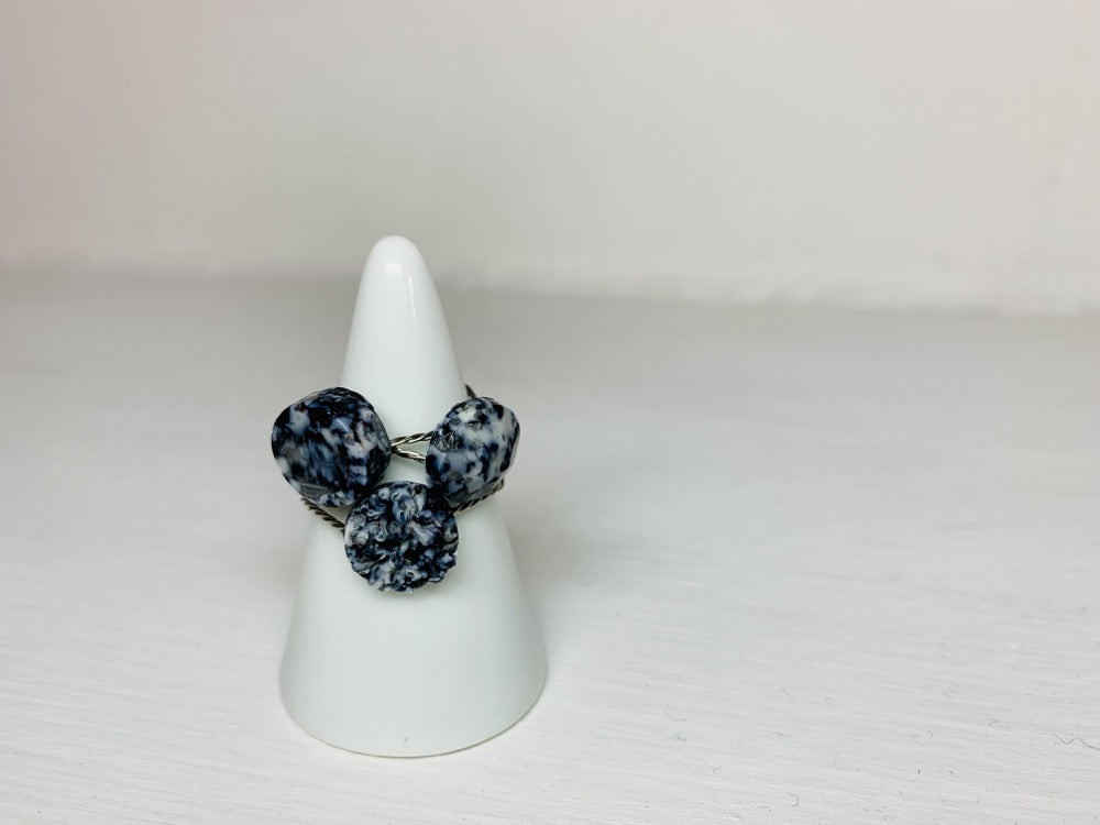 On a white background and slipped on a white cone ring holder are three rings cast from recycled 3D prints. The rings are each in a circular shape. One has a smooth surface, another a faceted surface, and finally one like a geode forming. They each have black, white, and silver colors intermixed to be speckled like granite.