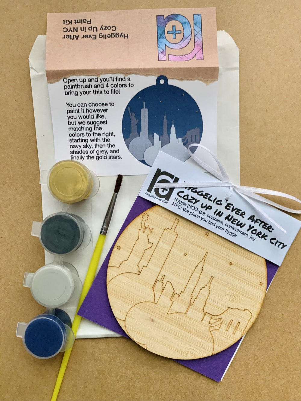 Shown laying on craft paper is the packaging and contents of this DIY Paint Kit. There is a paper envelope with instructions, a paintbrush, a set of 4 paint wells and a wall hanging with a laser cut NYC landmark.