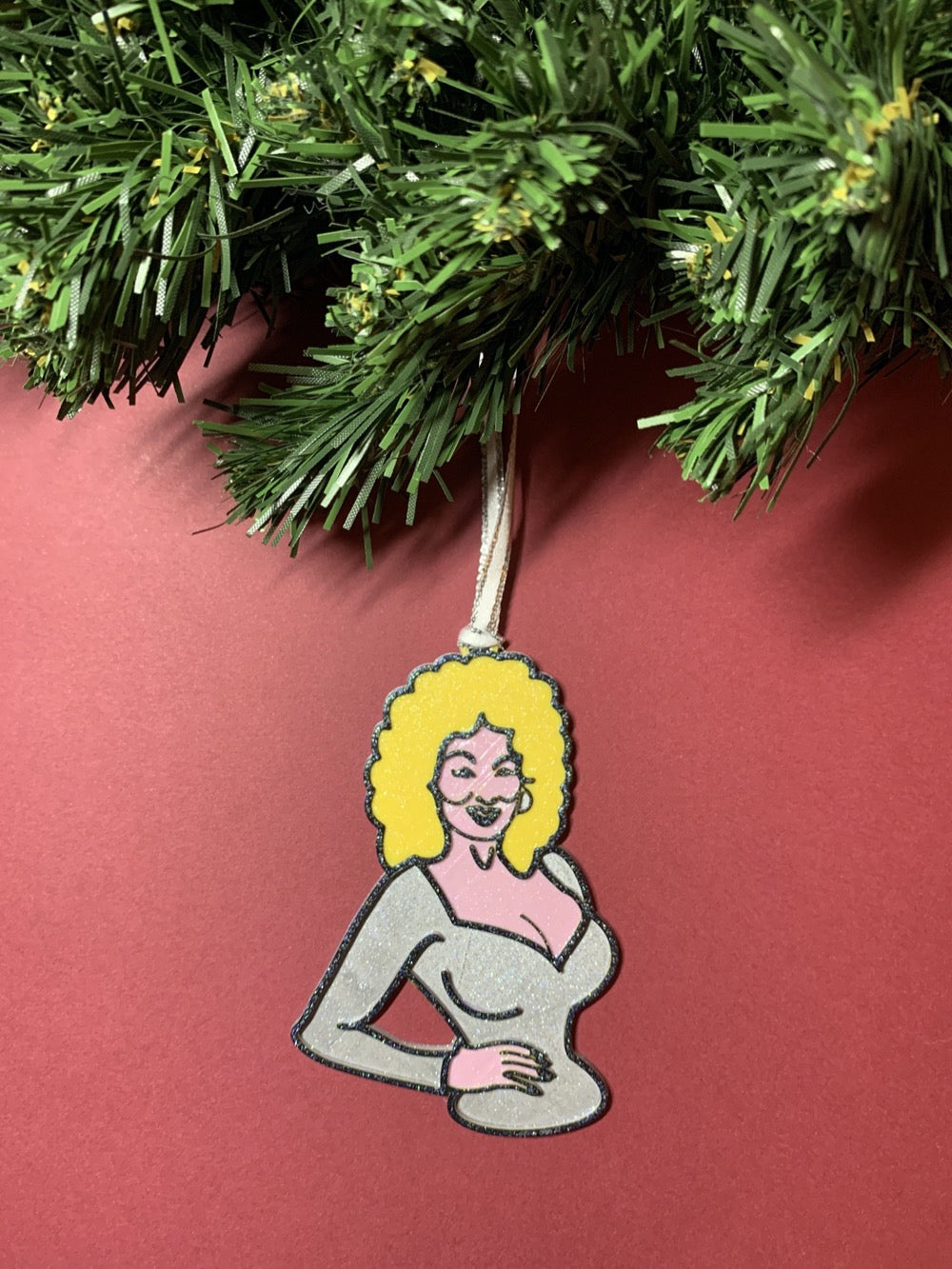 On a red background with a green wreath above is a R+D 3D printed ornament. This ornament is shaped like Dolly Parton. You can see one hand resting on her hip as she wears a silver top. Her trademark neckline and big blond hair is also visible with a smile. The entire ornament is covered in glitter to be able to shimmer and shine in the light.