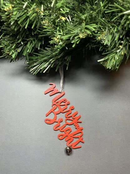 On a grey background and hanging from a green wreath is a 3D printed R+D ornament. It is a cursive text with a jingle bell and covered in glitter to make it shimmer and shine in the light. This ornament reads, Merry and Bright.