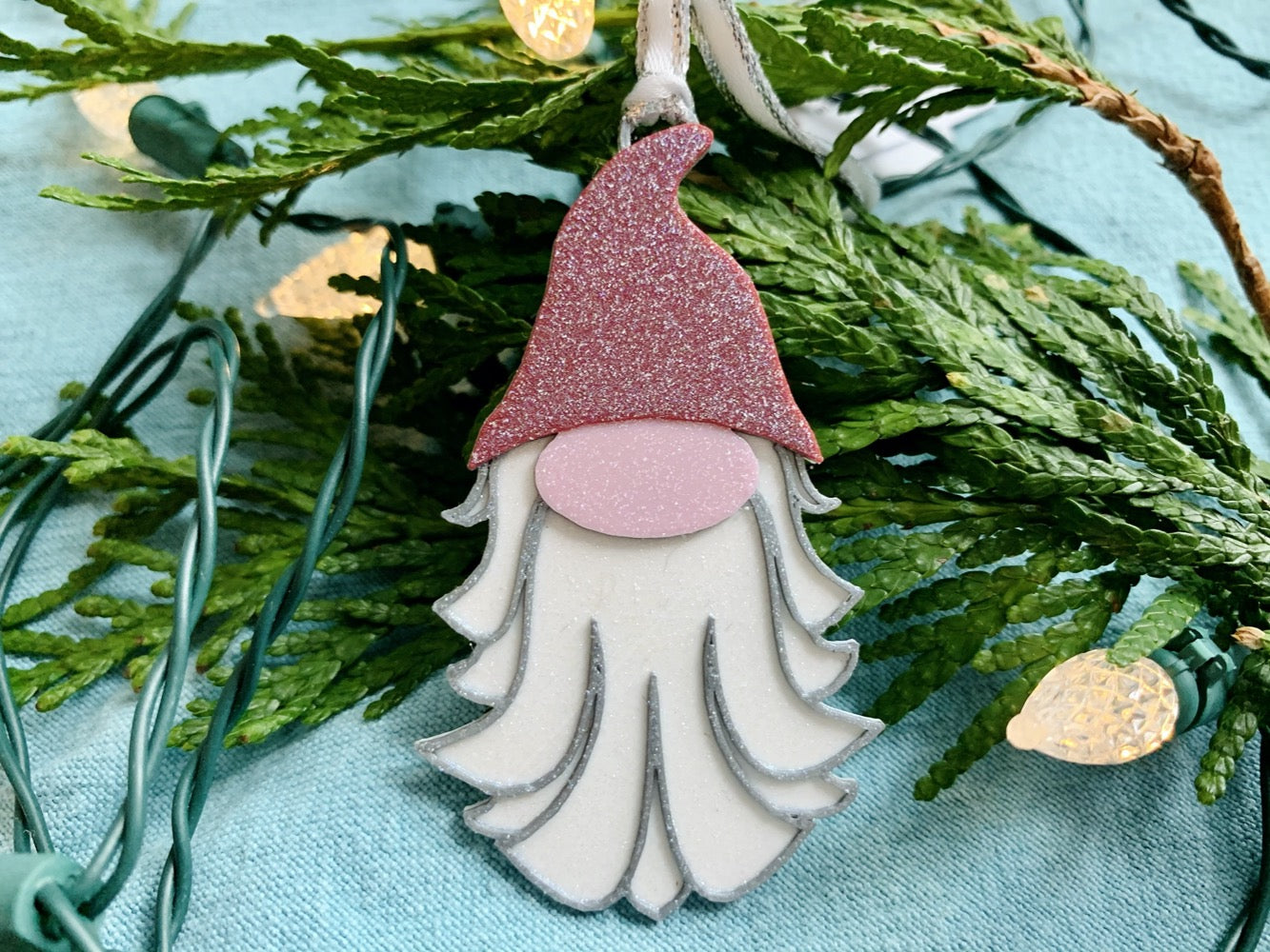 On a bright blue fabric background there are evergreen branches and a string of lights. Resting on these is a R+D 3D printed ornament. This ornament is shaped like a gnome, elf, or plump santa. It has a red pointed hat, a big pink nose, and a white and silver beard. The entire ornament is covered in glitter to be able to shimmer and shine in the light.