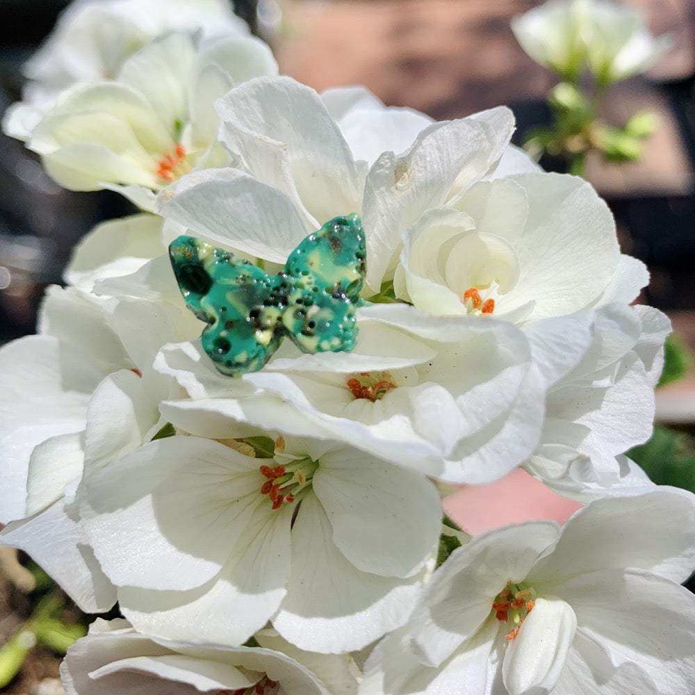 Resting in bright white geranium blooms is a ring that is shaped like a butterfly and cast from recycled 3D prints. The colors are bright hues of green and black. 