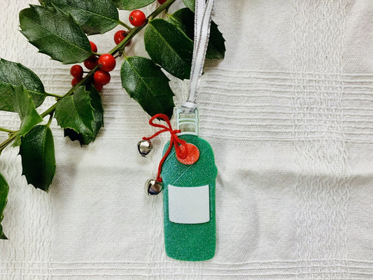 Shown on a white fabric background with a sprig of holly is a 3D printed R+D ornament.  It is in the shape of a gin bottle: green with a white label and a red emblem. Tied around the neck is a red bow that has two jingle bells attached. The entire ornament is covered in glitter to give it a shimmer and shine. 