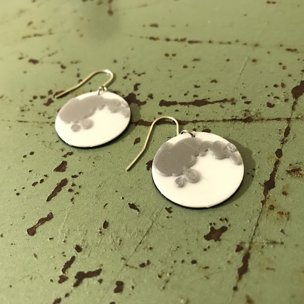 On a rusted and vintage green metal surface are two 3D printed earrings. They are both full moons with bright white plant based filament and silver filament to show the craters on the surface of the moon. 