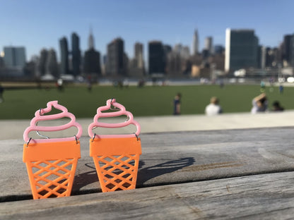 Two ice cream cone shaped earrings are perched between slats of a wooden bench. The top is pink and looks like soft serve swirled into a orange sugar cone. In the background, you can see a park with people enjoying the sun and the Manhattan skyline. 