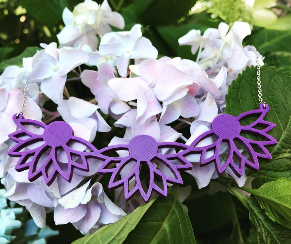 Hanging from a  hydrangea bloom is a 3D printed necklace. It is a bib necklace that stretches to have three daisys with petals stretching downward. This necklace is printed in a sustainable purple filament.