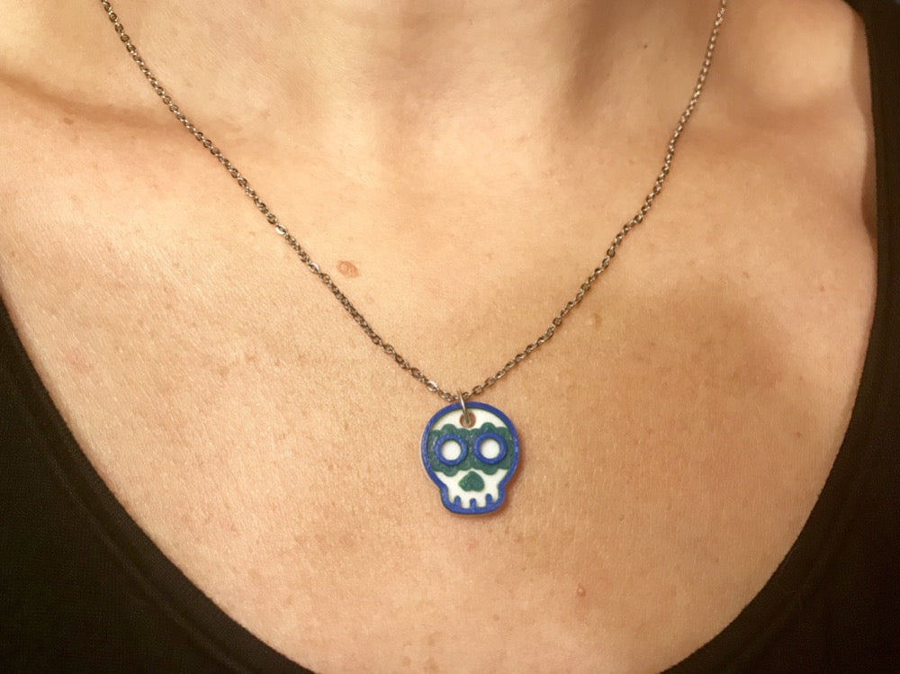 A necklace is being shown worn around someone's neck. There is a thin chain and a 3D printed pendant. The pendant is a small sugar skull that is white, green, and blue. 